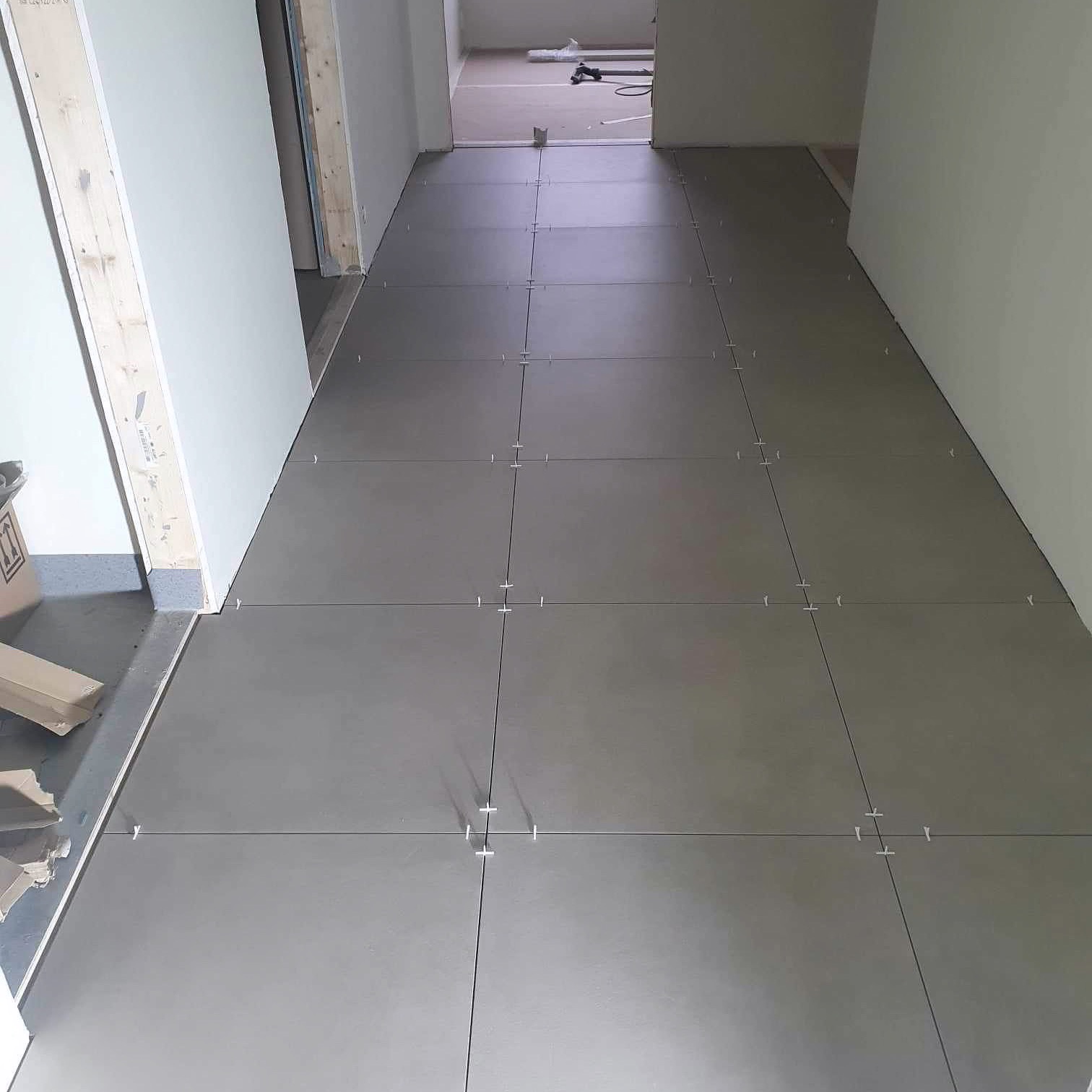 Układanie szarych płytek podłogowych w korytarzu, z widocznymi krzyżykami dystansowymi między płytkami.
