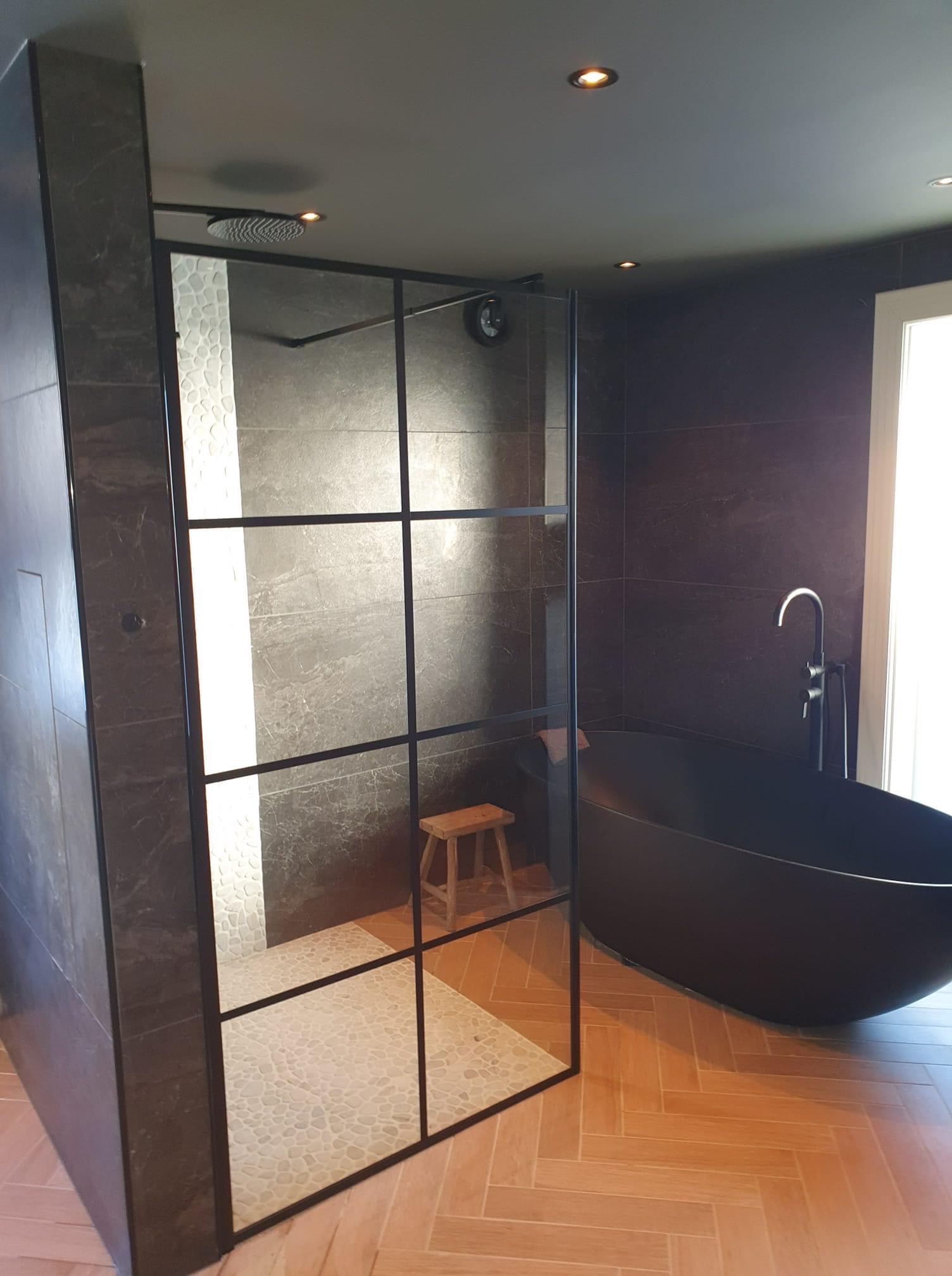 Nowoczesna łazienka z czarną wanną wolnostojącą i kabiną prysznicową typu walk-in z czarnymi profilami, wykończona płytkami w odcieniach szarości i drewnianą podłogą ułożoną w jodełkę.