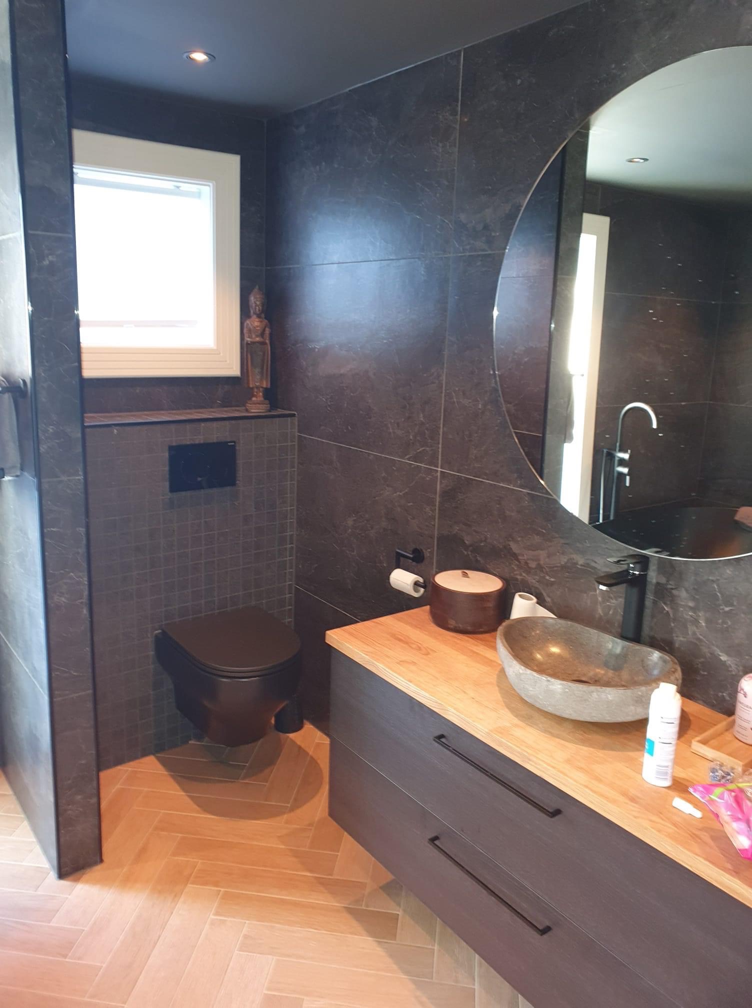 Nowoczesna łazienka z ciemnymi płytkami imitującymi kamień, drewnianą podłogą w jodełkę, owalną umywalką z kamienia i okrągłym lustrem bez ramy.