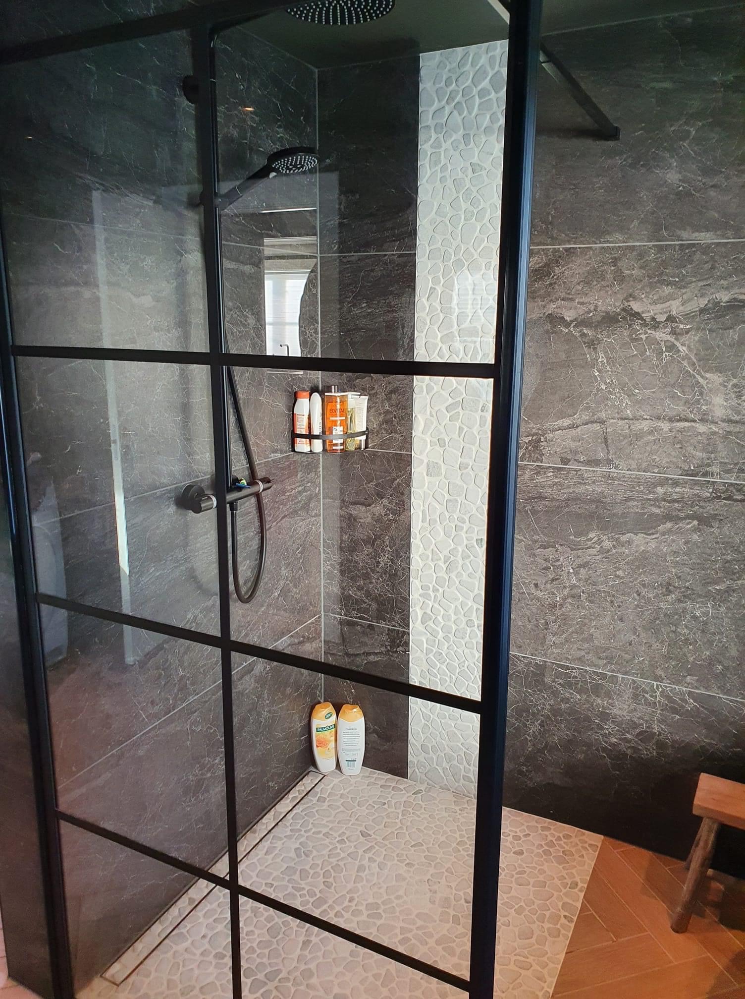 Nowoczesna kabina prysznicowa z czarną ramą, marmurowymi płytkami i mozaiką z kamyków, uzupełniona o drewniany stołek i kosmetyki w koszyku.