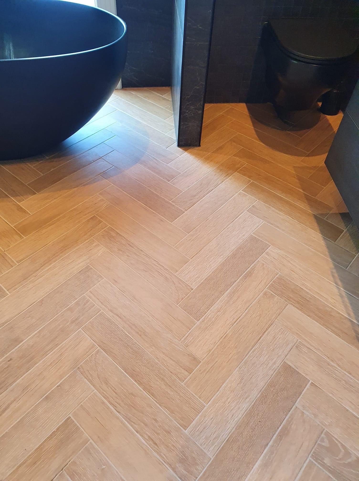 Jasnobrązowa podłoga z płytek ułożonych w jodełkę w nowoczesnej łazience z czarną wanną wolnostojącą i toaletą.