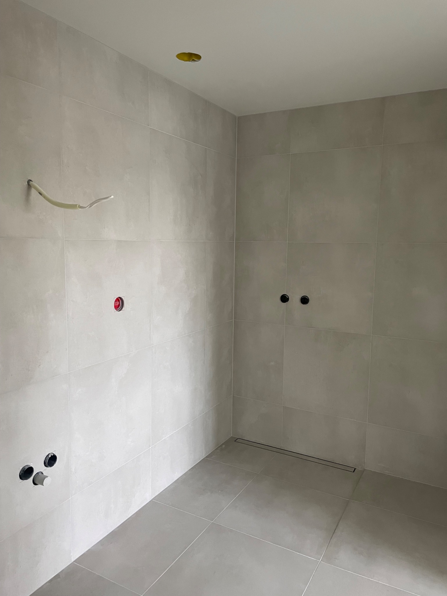 Surowe wnętrze łazienki w szarych płytkach, widoczne przygotowane instalacje wodno-kanalizacyjne i elektryczne, odpływ liniowy w podłodze.