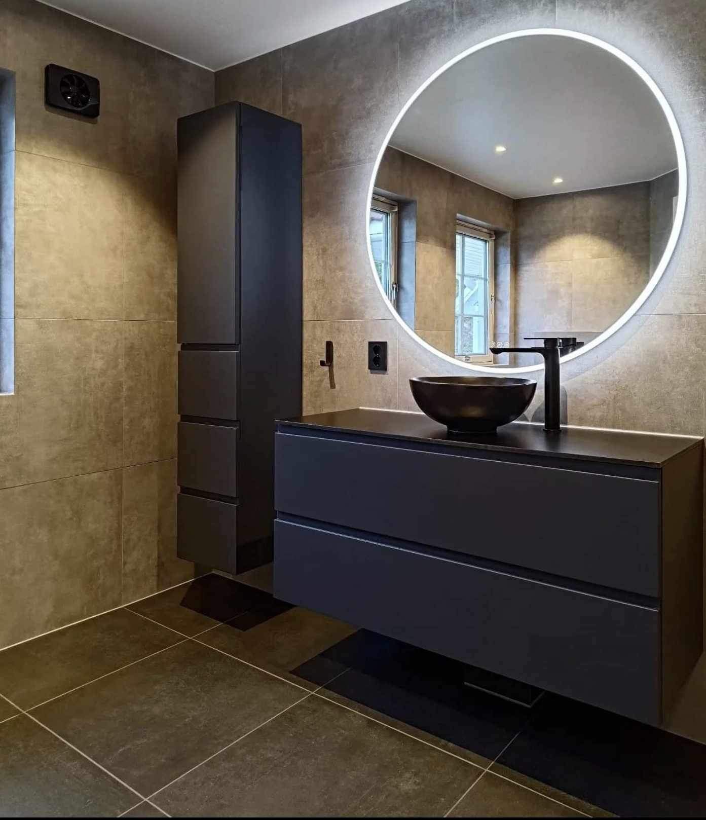 Nowoczesna łazienka z ciemnoszarą armaturą, okrągłym lustrem z podświetleniem LED i płytkami imitującymi beton.