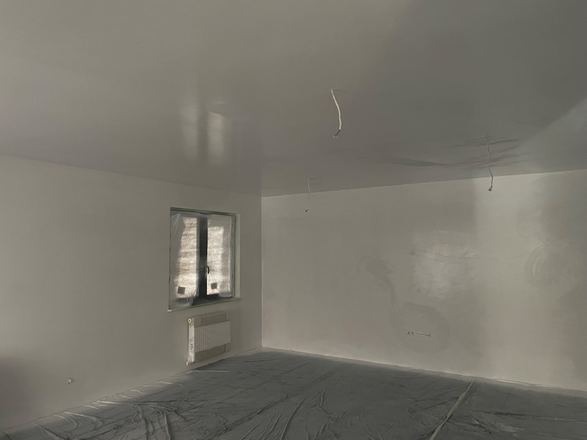 Pokój w trakcie prac wykończeniowych: świeżo pomalowane na biało ściany i sufit, zabezpieczona folią okno i podłoga, wystające kable elektryczne.