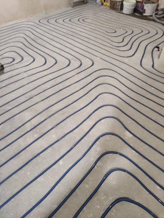 Niebieskie rurki ogrzewania podłogowego ułożone w zygzak na surowym, betonowym podłożu przed wylaniem wylewki.