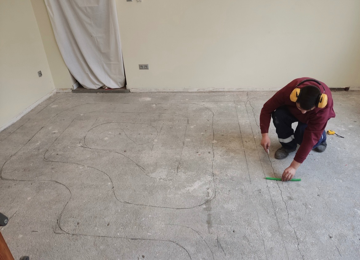 Pracownik w słuchawkach rysuje ołówkiem linie na betonowej posadzce, wyznaczając trasę pod przyszłe rury grzewcze, używając zielonej listwy jako linijki.