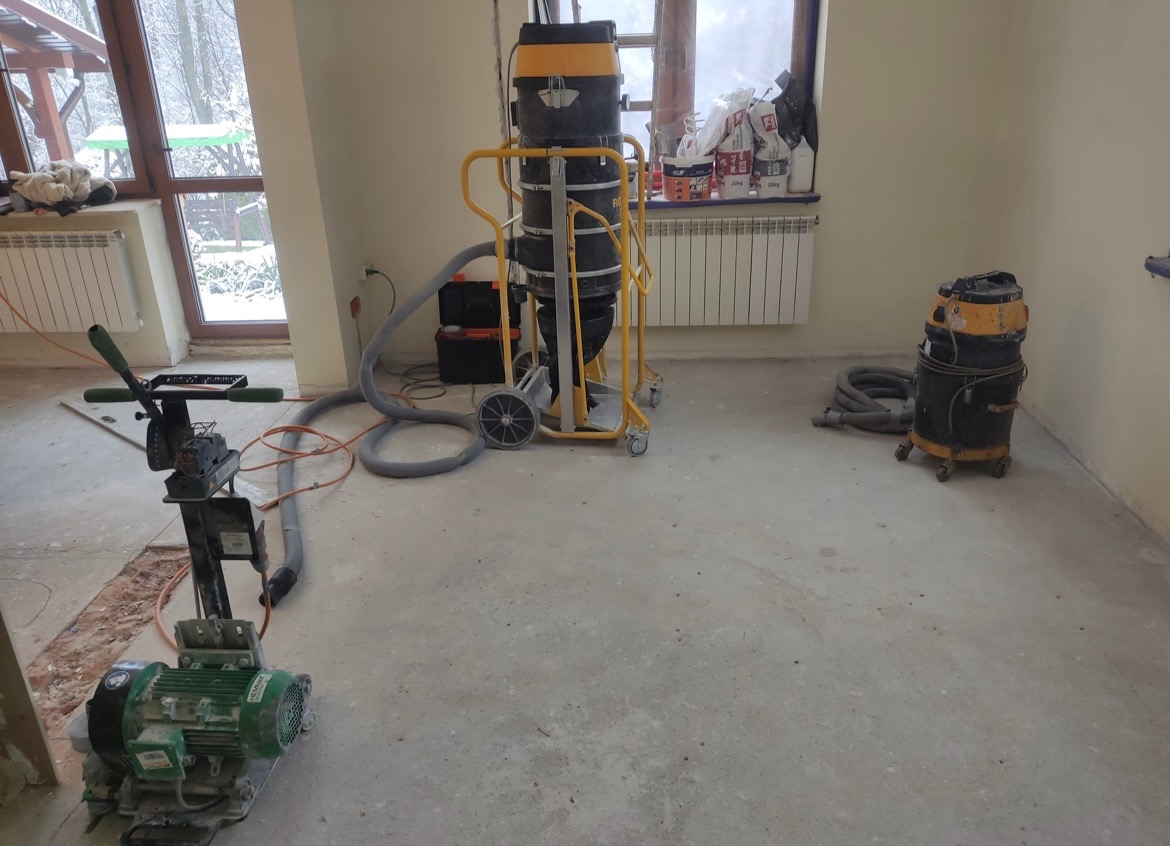 Przygotowanie podłoża pod ogrzewanie podłogowe: szlifierka do betonu z podłączonym odkurzaczem przemysłowym na kółkach w pomieszczeniu z oknem i grzejnikiem.