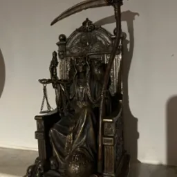 Brązowa statuetka Santa Muerte siedzącej na tronie, trzymającej wagę i kosę, z czaszkami u podstawy, umieszczona na marmurowym blacie pod białą ścianą.