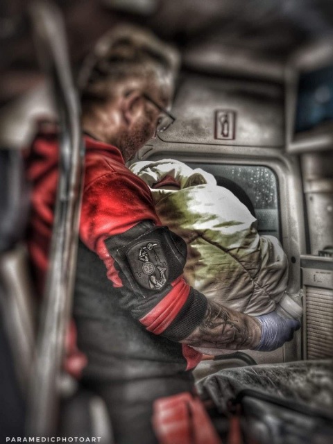 Ratownik medyczny w czerwonej bluzie z naszywką i tatuażem na ramieniu, trzymający zawinięte dziecko w ambulansie