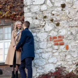 Uśmiechnięta para trzymająca się za ręce na tle muru z białego kamienia i cegieł, kobieta w beżowym płaszczu i szaliku, mężczyzna w granatowym płaszczu.