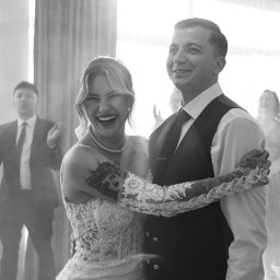 Sobieraj Photo - Czarno-białe zdjęcie uśmiechniętej panny młodej z tatuażem na ramieniu i pana młodego podczas pierwszego tańca, z gośćmi w tle.