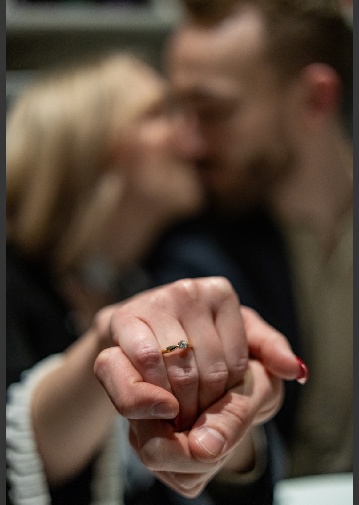 Zaręczynowy pierścionek z brylantem na dłoni kobiety, w tle całująca się para, ujęcie z bliska.