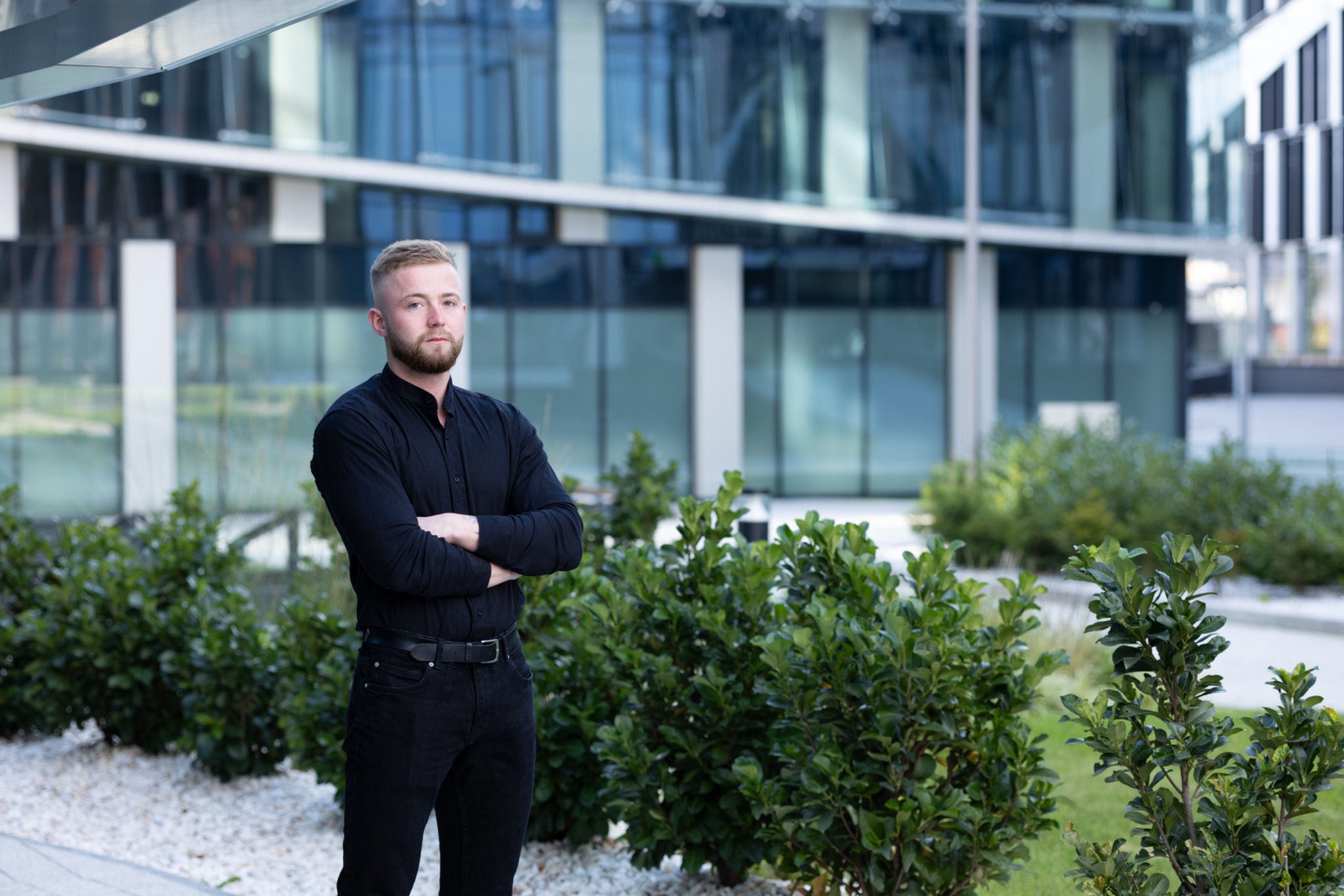 Młody mężczyzna w czarnym ubraniu stoi z założonymi rękami na tle nowoczesnego biurowca i zielonych krzewów.