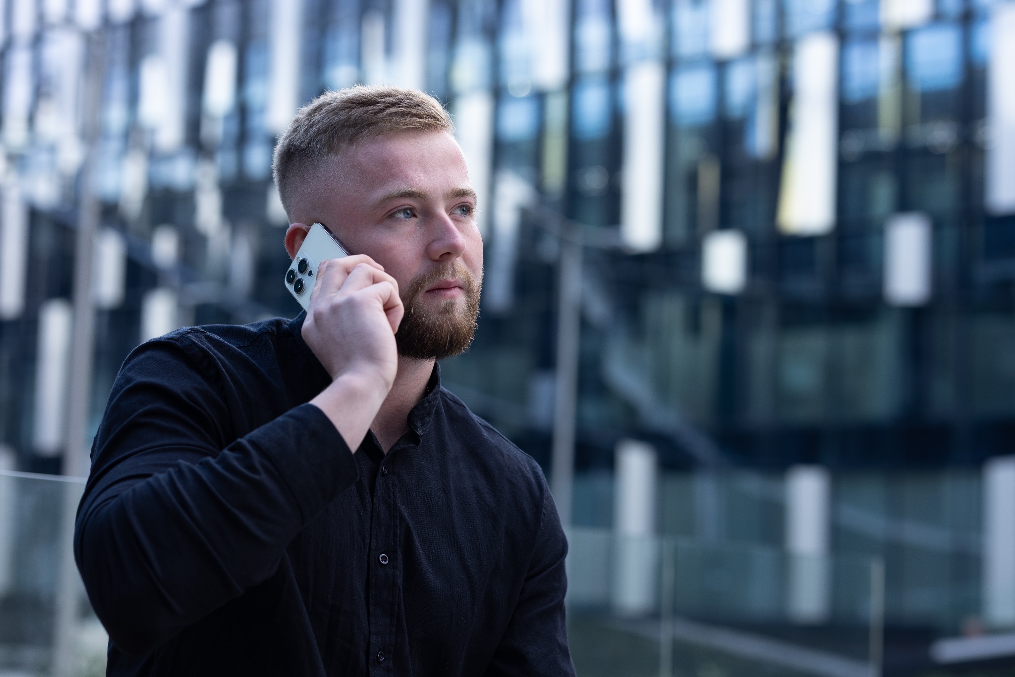 Młody mężczyzna z brodą w czarnej koszuli rozmawia przez telefon na tle szklanego budynku biurowego.