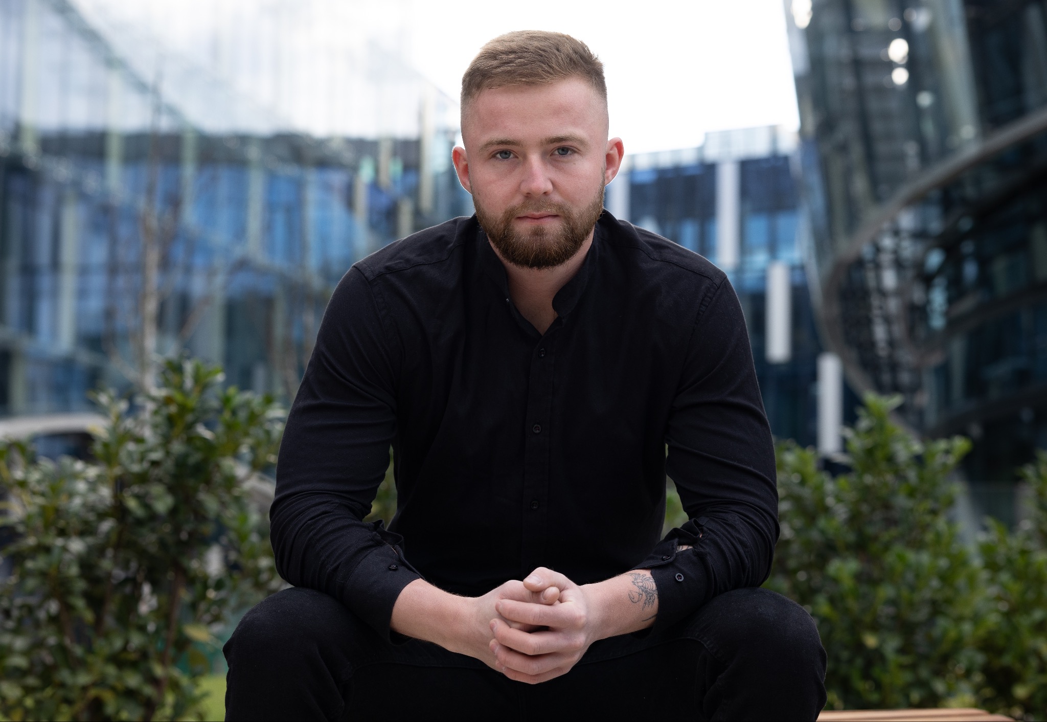 Portret młodego mężczyzny z brodą w czarnej koszuli, z tatuażem na przedramieniu, siedzącego na tle nowoczesnego budynku biurowego.