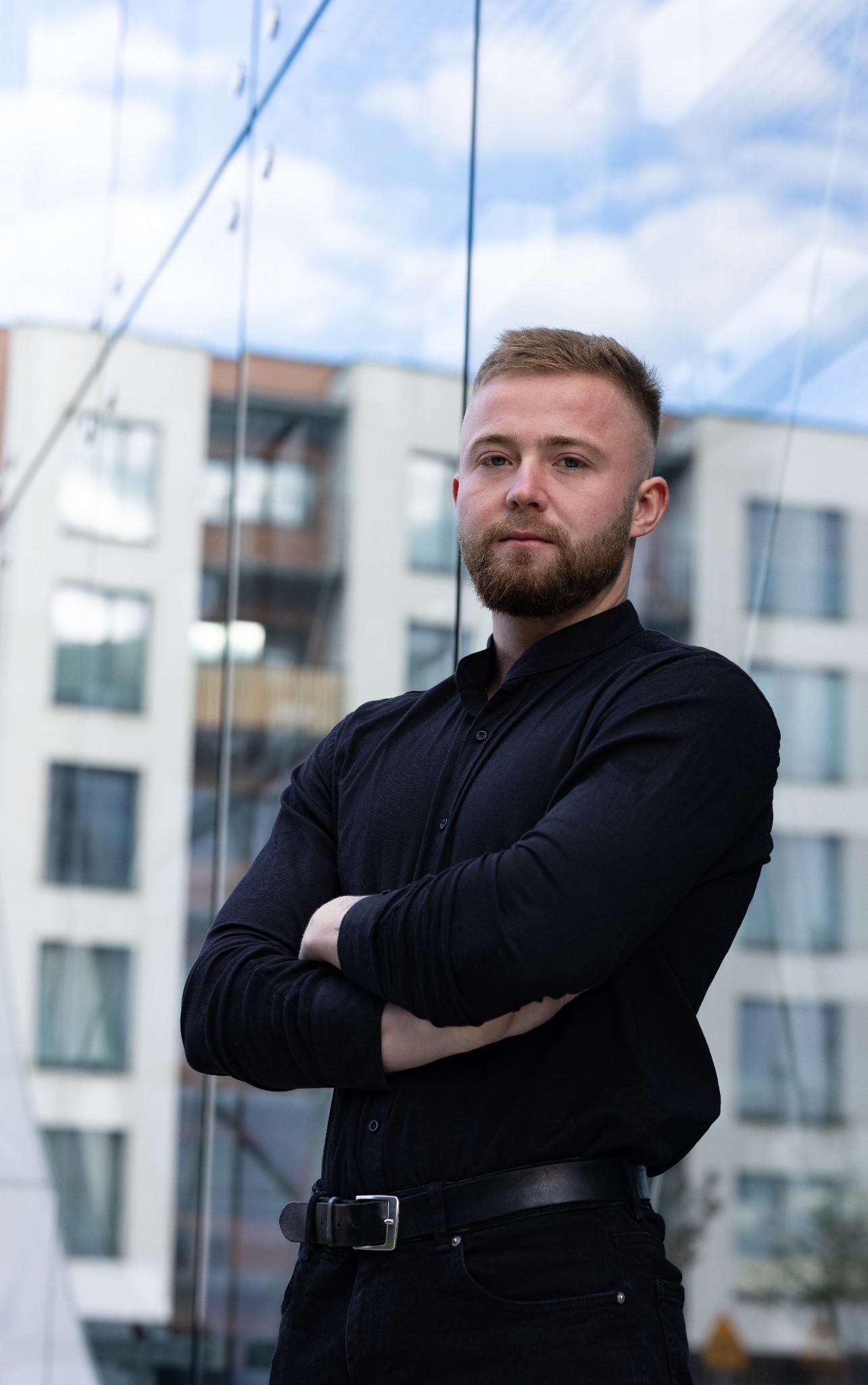 Portret mężczyzny z brodą w czarnej koszuli, z założonymi rękami, na tle szklanej fasady budynku.