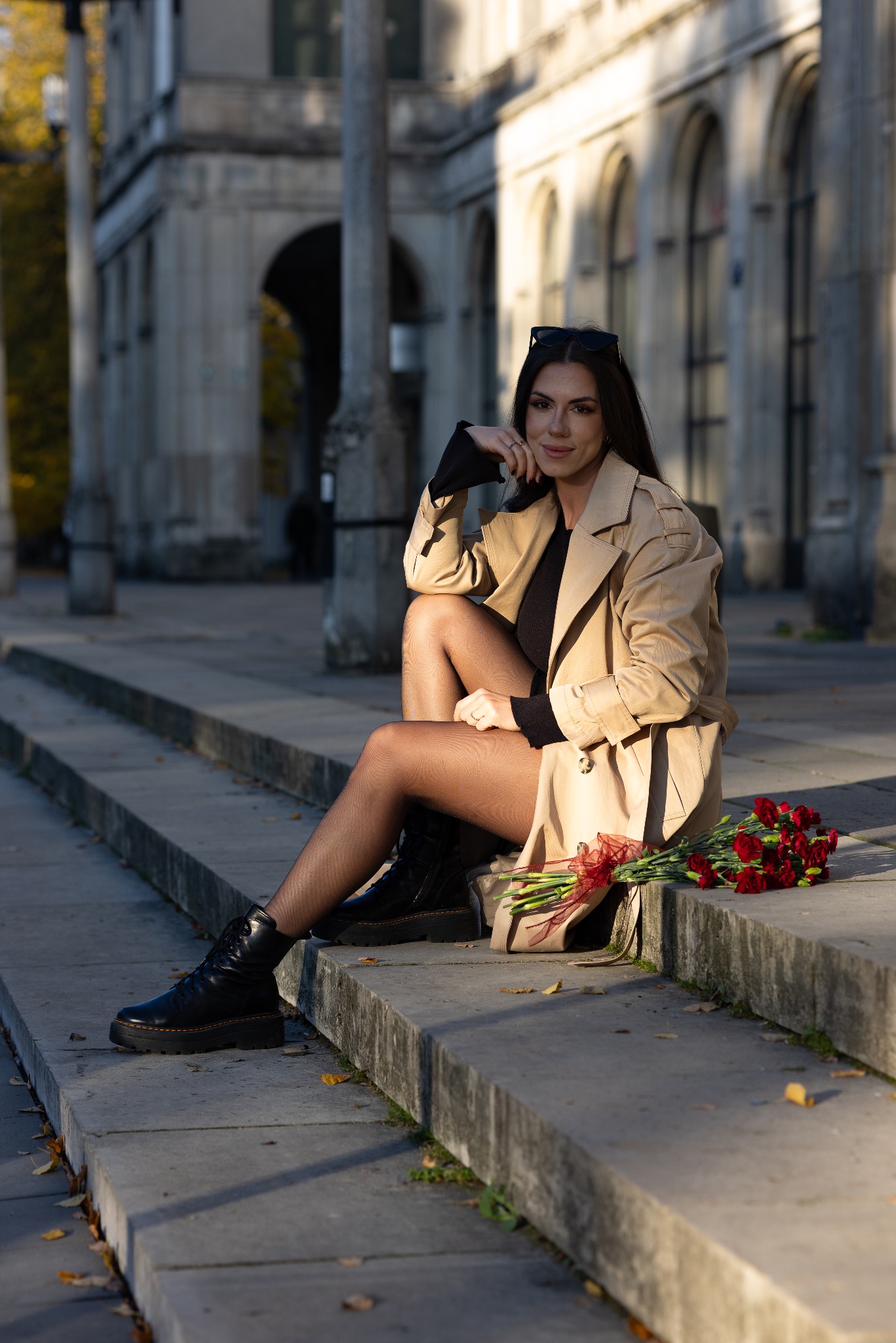 Kobieta w beżowym trenczu i czarnych botkach siedzi na betonowych schodach, obok leży bukiet czerwonych goździków z czerwoną wstążką, w tle widoczny budynek z kolumnami.
