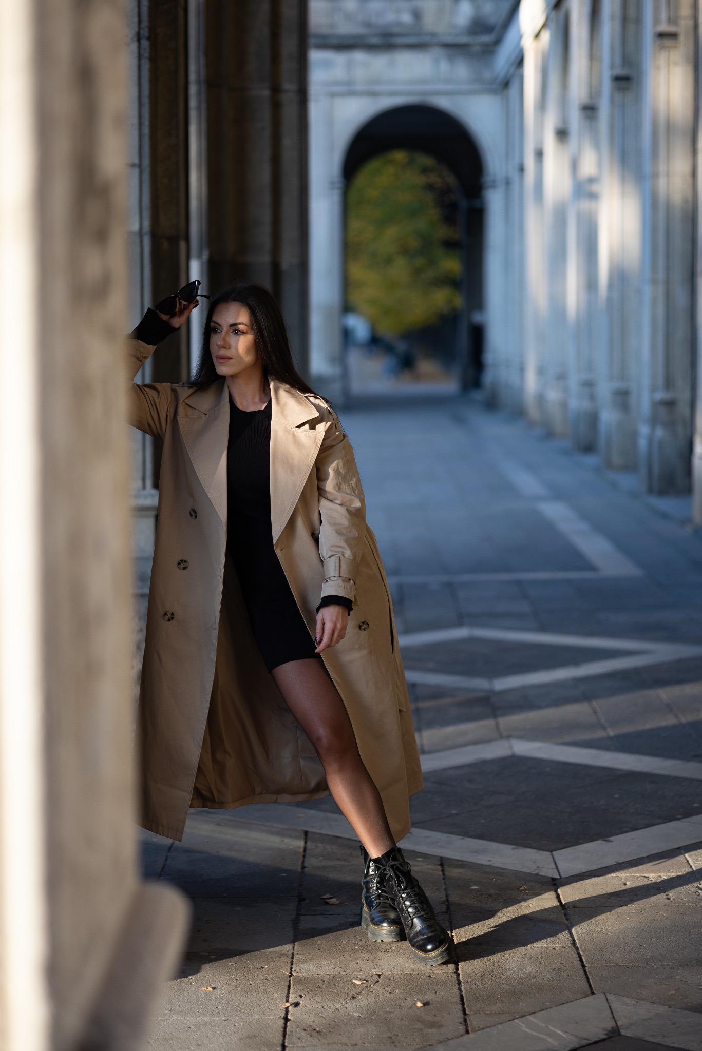 Kobieta w beżowym trenczu i czarnej sukience pozuje w architektonicznej przestrzeni z kolumnami i łukami.