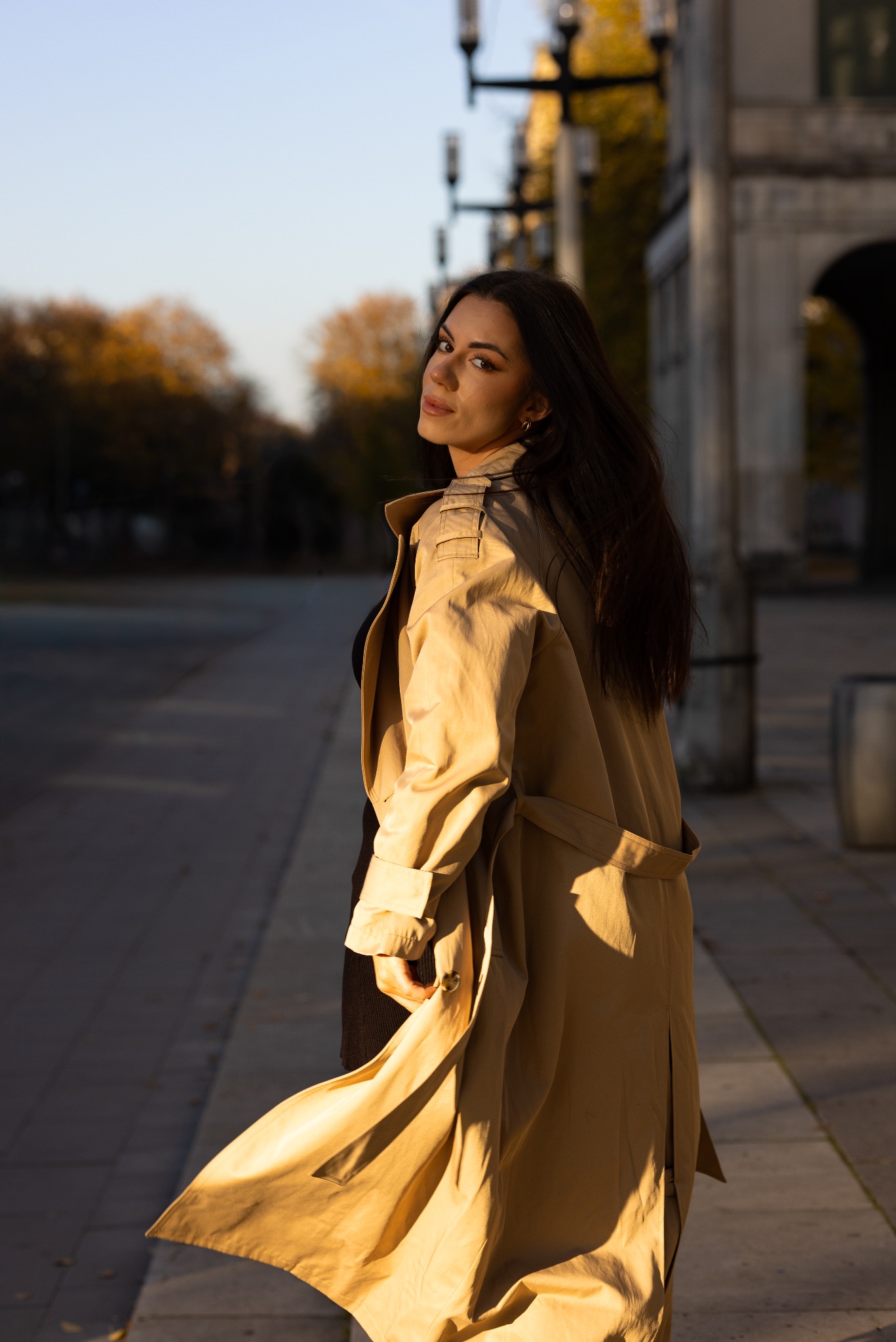 Kobieta w beżowym trenczu odwraca się, patrząc przez ramię na tle miejskiego krajobrazu o zachodzie słońca.