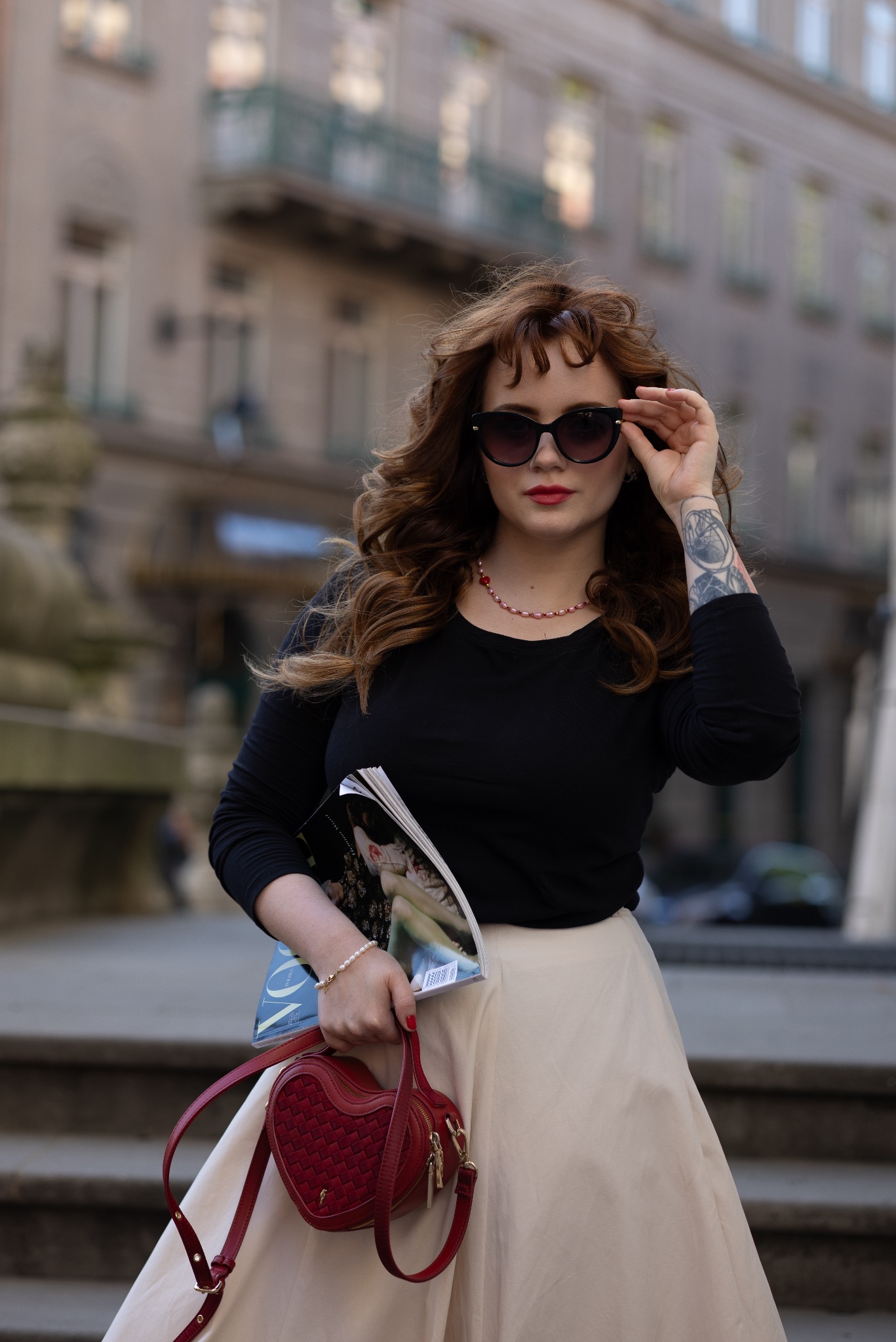 Kobieta w okularach przeciwsłonecznych, czarnej bluzce i beżowej spódnicy trzyma w dłoniach magazyn i czerwoną torebkę w kształcie serca, na tle miejskiej architektury.