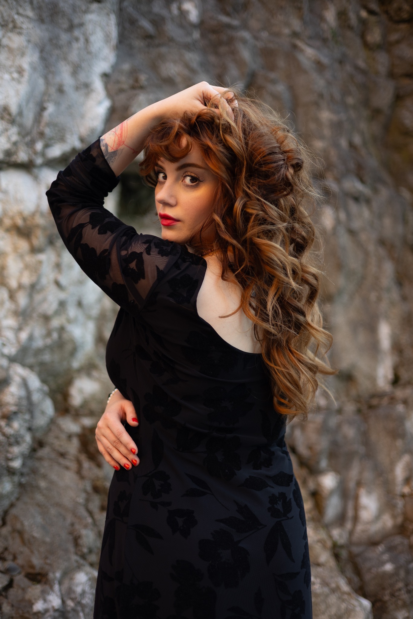 Kobieta w czarnej sukience z kwiatowym wzorem, pozująca na tle skalnej ściany, z tatuażem na ramieniu i czerwonymi ustami.