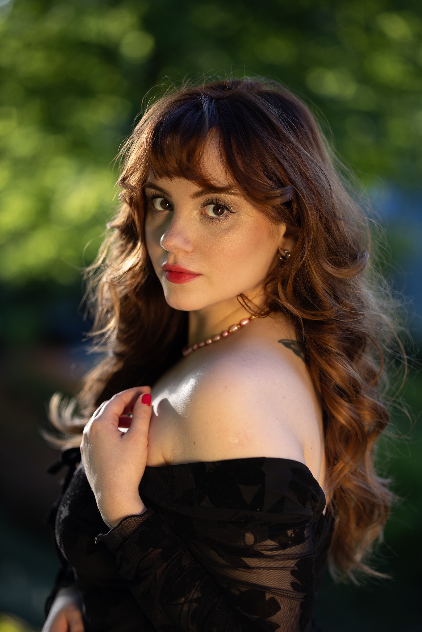 Portret kobiety z rudymi włosami i czerwonymi ustami, w czarnej sukience z odkrytymi ramionami, z delikatnym naszyjnikiem z pereł i tatuażem na ramieniu, pozującej na tle zieleni.