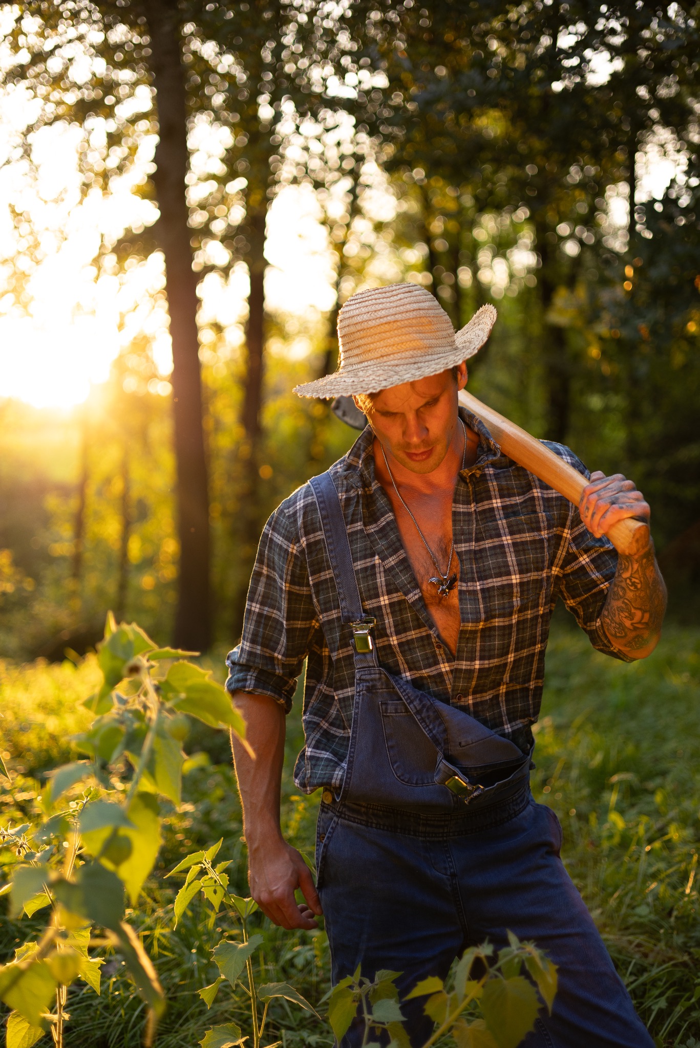 Mężczyzna w słomkowym kapeluszu i dżinsowych ogrodniczkach, niosący siekierę na ramieniu w zalesionym terenie oświetlonym zachodzącym słońcem.