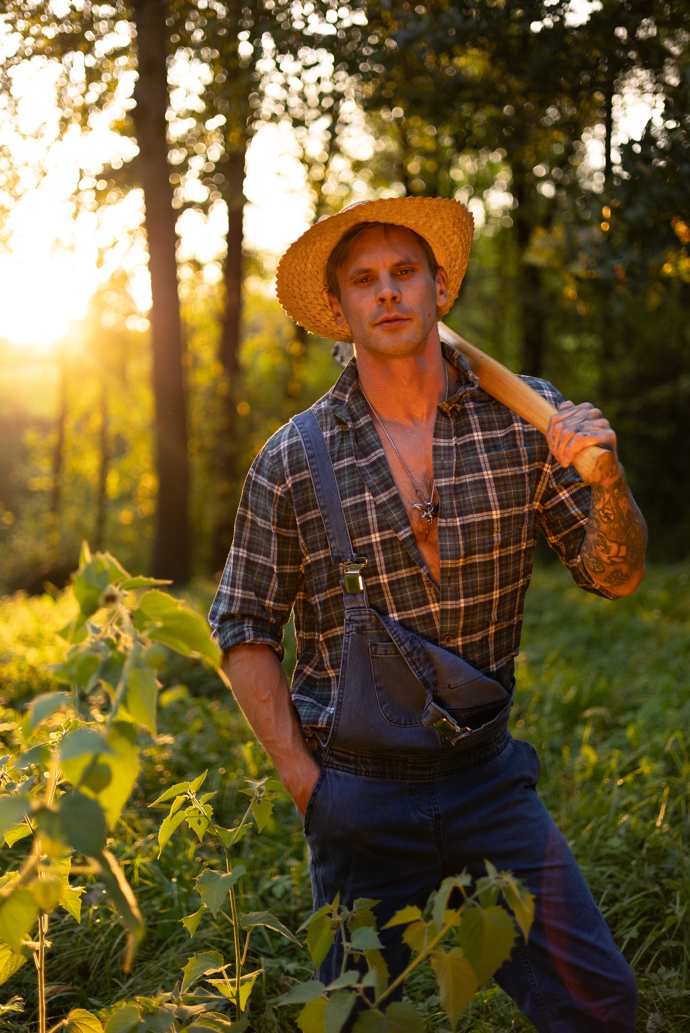 Mężczyzna w słomkowym kapeluszu, kraciastej koszuli i ogrodniczkach, z siekierą na ramieniu, stoi w zaroślach w promieniach zachodzącego słońca. Widoczny tatuaż na ramieniu i naszyjnik z zawieszką.