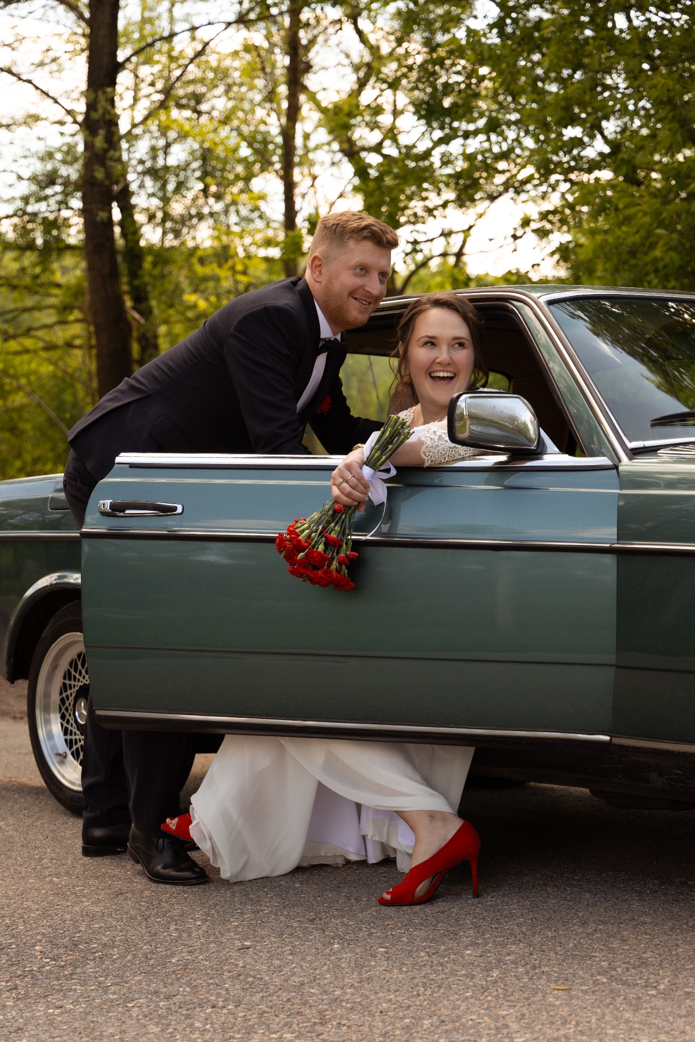 Uśmiechnięta para młoda w zielonym, klasycznym samochodzie. Panna młoda w czerwonych szpilkach trzyma bukiet czerwonych goździków. Romantyczna sesja w plenerze.