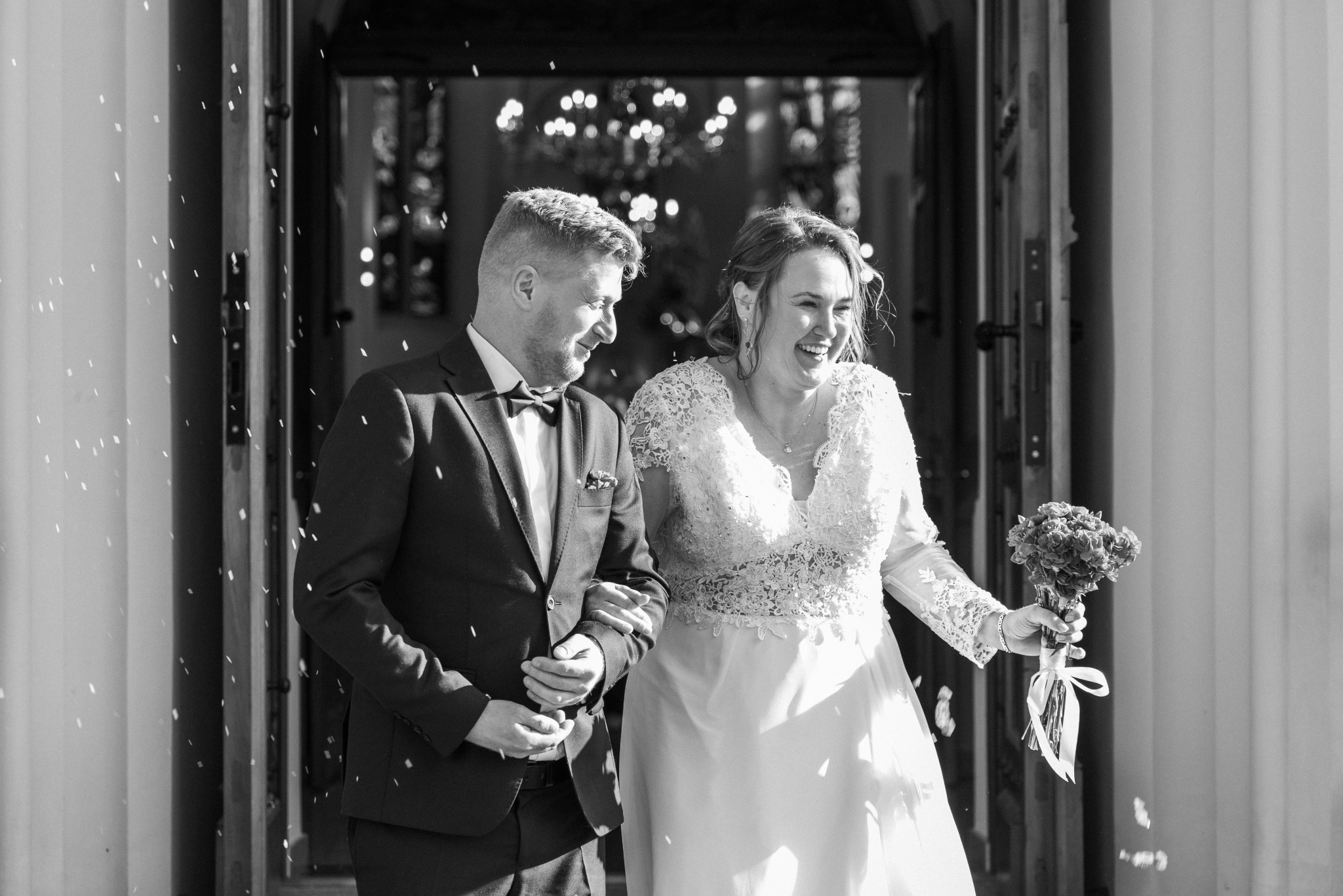 Czarno-białe zdjęcie uśmiechniętej pary młodej wychodzącej z kościoła, obsypywanej konfetti. Pan młody w smokingu, panna młoda z bukietem. Detal: widoczne zdobienia drzwi.
