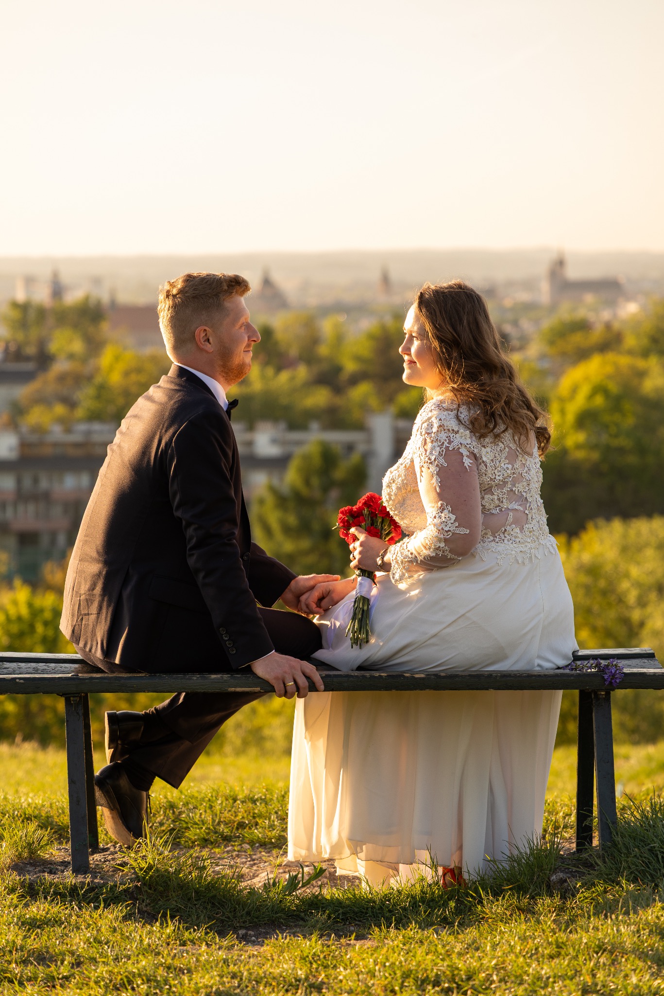 Para młoda siedzi na ławce na tle panoramy miasta w promieniach zachodzącego słońca. Panna młoda trzyma bukiet czerwonych róż.