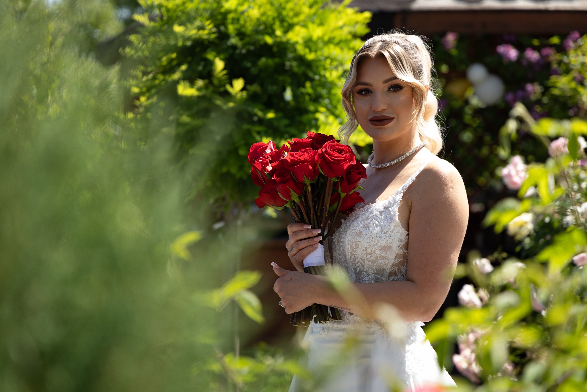 Panna młoda w białej sukni trzyma bukiet czerwonych róż na tle zieleni. Delikatny makijaż, biżuteria i upięte włosy dopełniają elegancki wygląd.