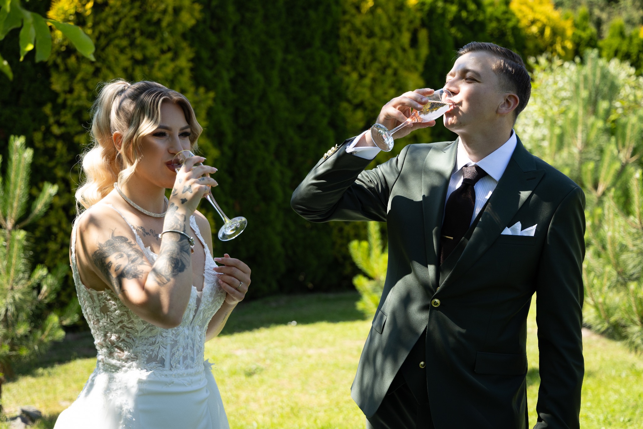 Para młoda pije szampana na tle zieleni. Panna młoda w sukni z koronkowym gorsetem, pan młody w eleganckim, ciemnozielonym garniturze. Słoneczny dzień.