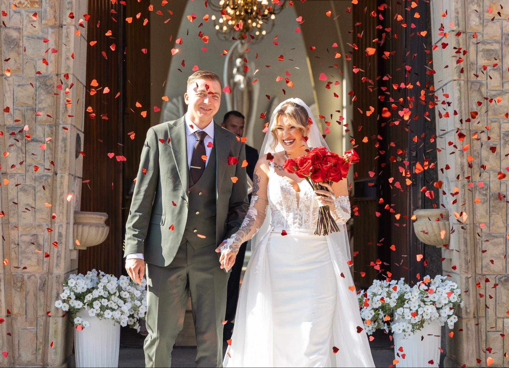 Uśmiechnięta para młoda wychodzi z kościoła, obsypana konfetti w kształcie serc. Panna młoda trzyma bukiet czerwonych róż, a w tle widać dekoracje kwiatowe.