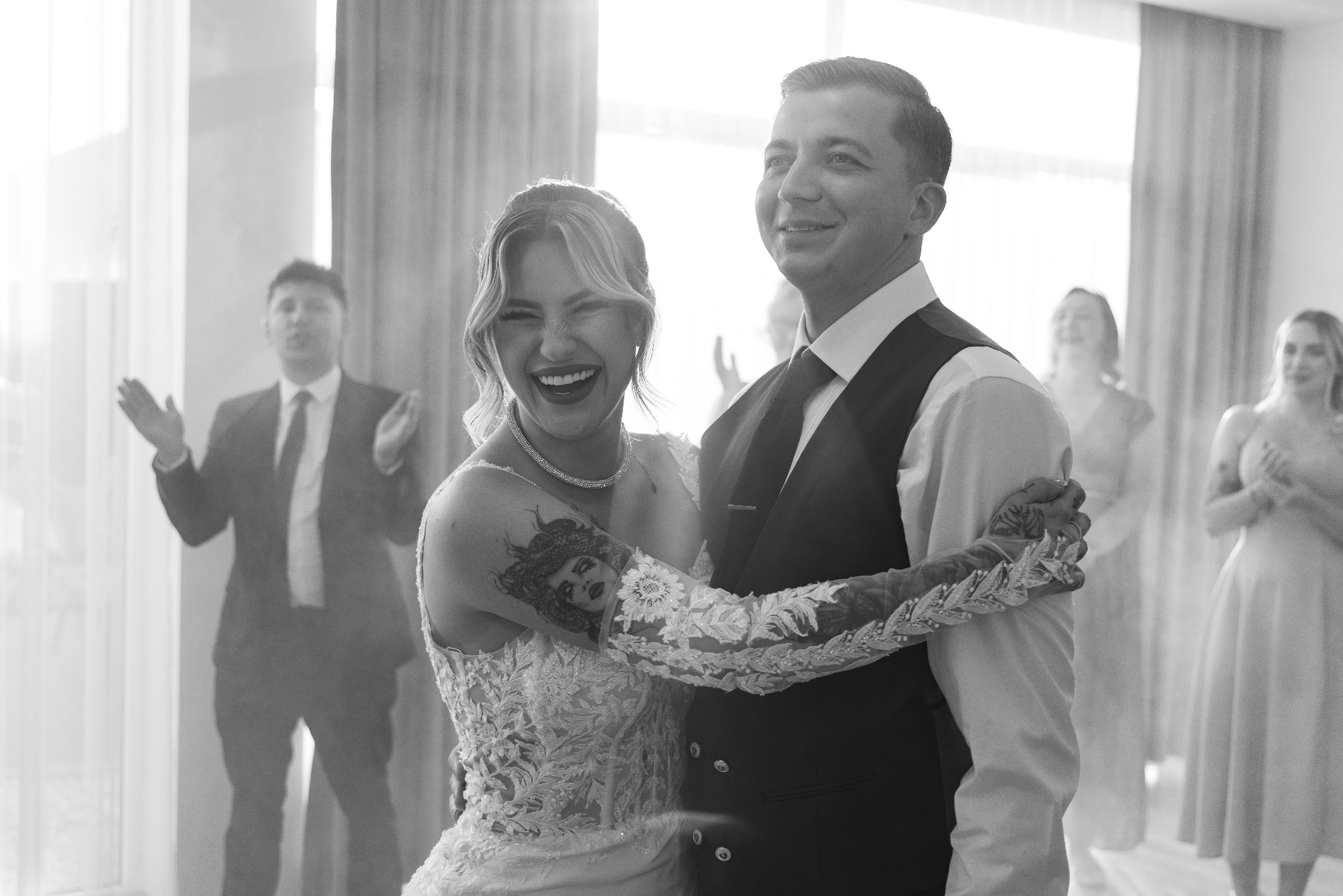 Czarno-białe zdjęcie uśmiechniętej panny młodej z tatuażem na ramieniu i pana młodego podczas pierwszego tańca, z gośćmi w tle.