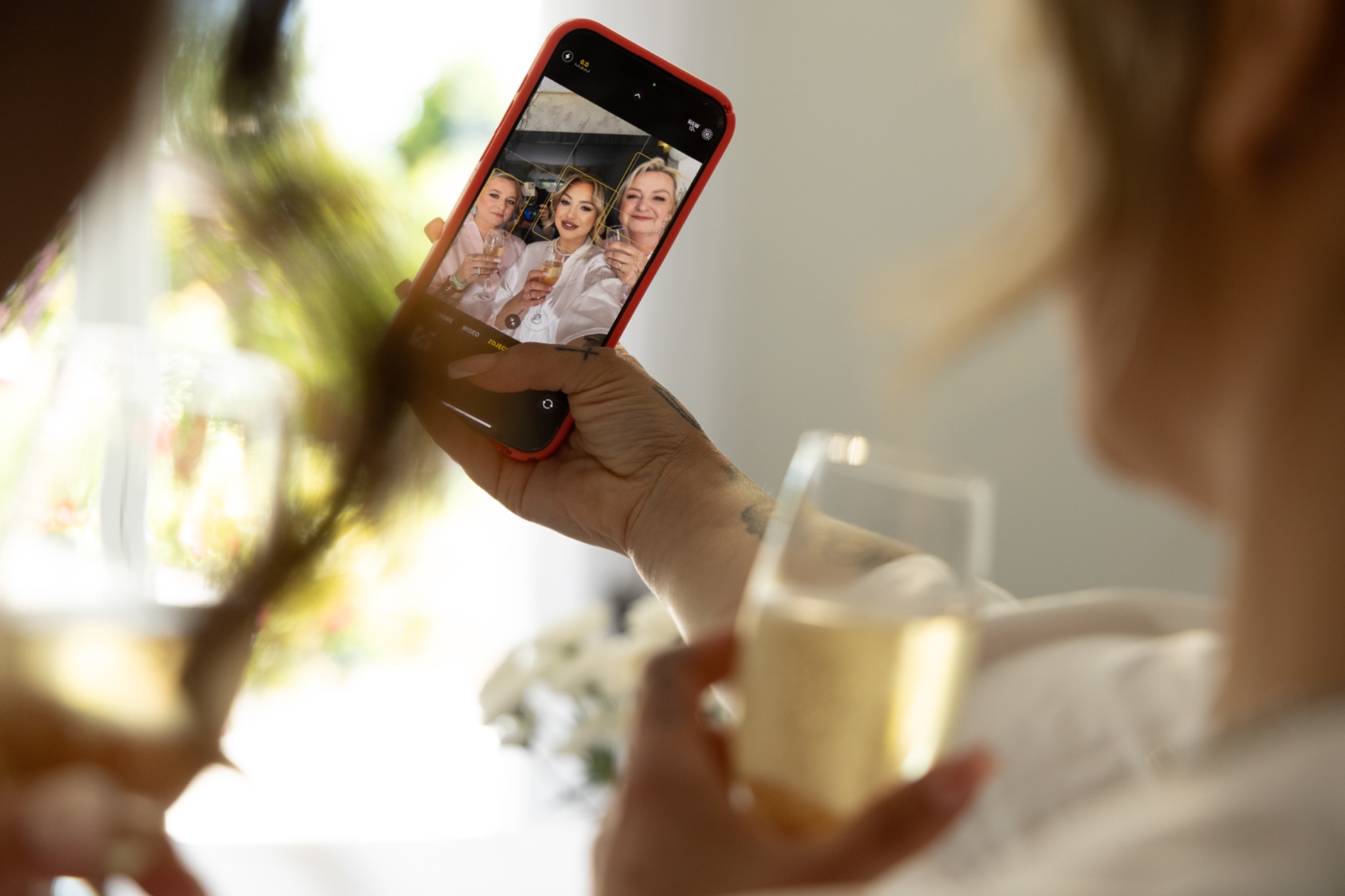 Kobieta trzyma telefon z widocznym zdjęciem trzech uśmiechniętych kobiet w szlafrokach, wznoszących toast szampanem. W tle rozmyte kwiaty i okno.