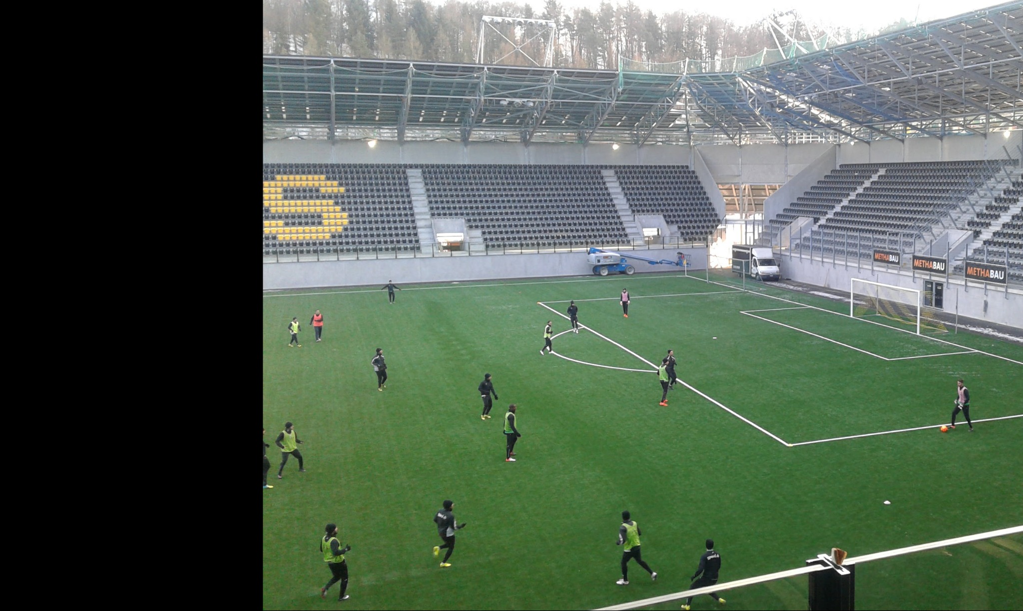 Widok z góry na boisko piłkarskie ze sztuczną murawą podczas treningu, z widocznymi trybunami, zadaszeniem konstrukcji stalowej oraz podnośnikiem koszowym i busem w tle.