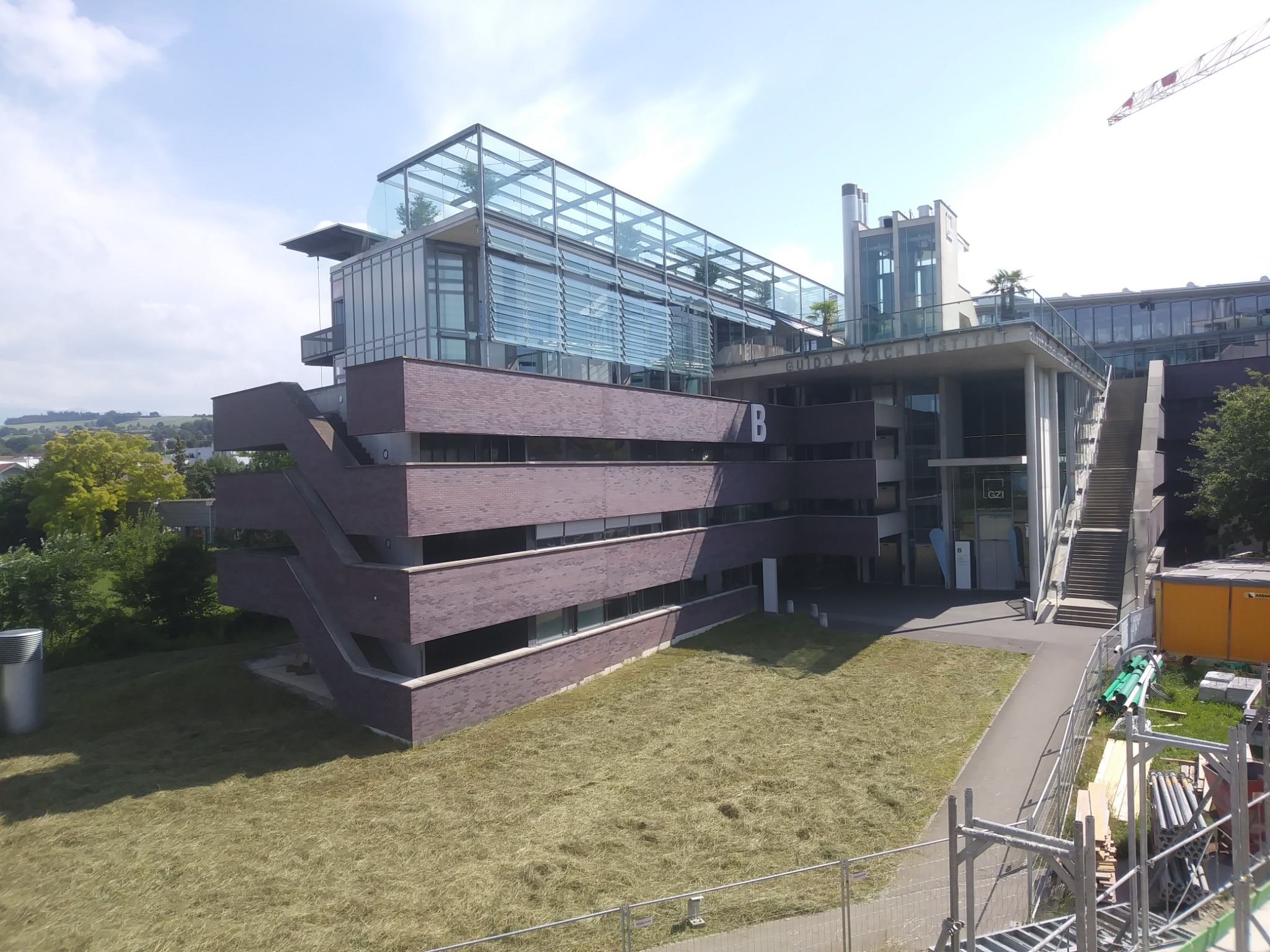 Nowoczesny budynek z cegły klinkierowej z przeszkloną nadbudówką i schodami zewnętrznymi, widok z góry na teren budowy z ogrodzeniem i składowiskiem materiałów.