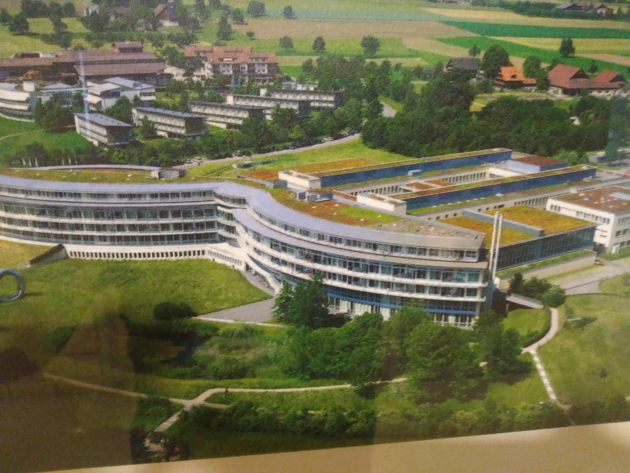 Widok z góry na kompleks budynków z zielonymi dachami, otoczony parkiem i polami uprawnymi w tle.