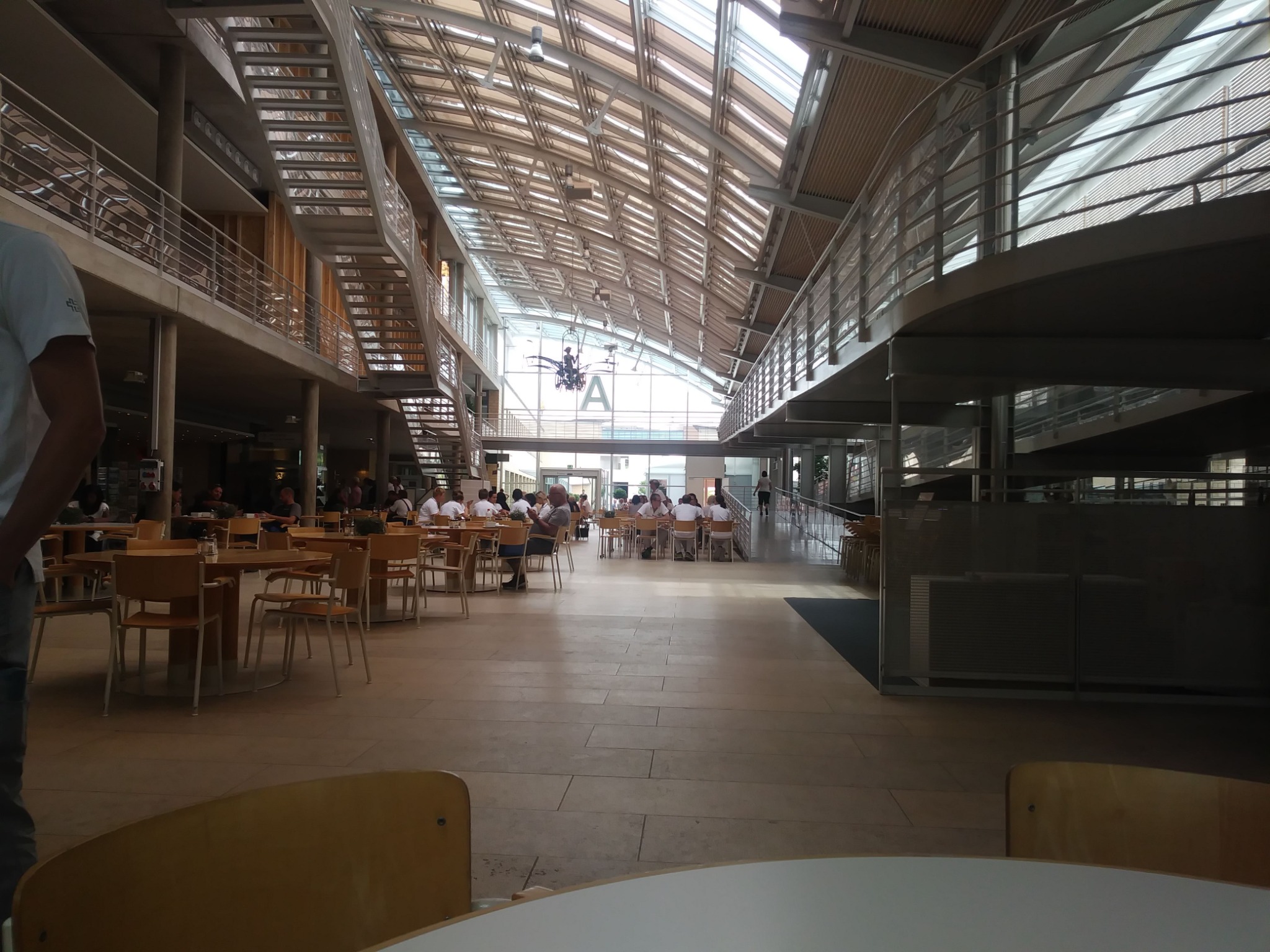 Przestronne wnętrze budynku z przeszklonym dachem, widoczne schody, balustrady i stoliki z krzesłami, oświetlone naturalnym światłem wpadającym przez dach.