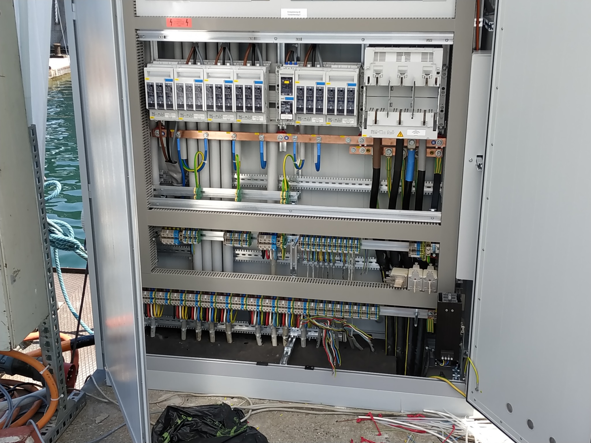 Otwarta szafa rozdzielcza z widocznymi bezpiecznikami, przewodami w różnych kolorach, szynami montażowymi i elementami łączeniowymi, z widokiem na wodę w tle.