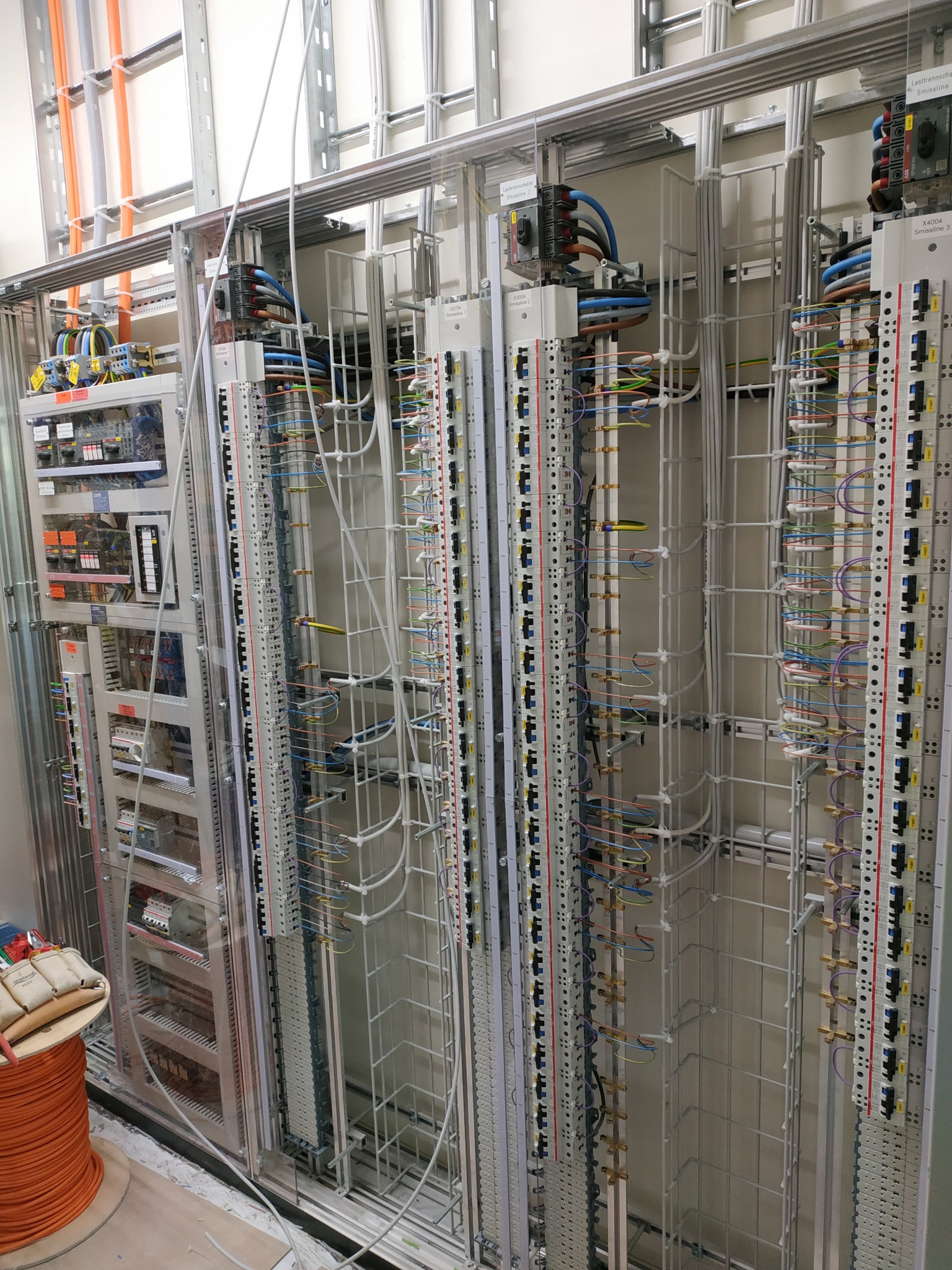 Rozbudowana szafa sterownicza z licznymi modułami, przewodami i listwami zaciskowymi; widoczny bęben z pomarańczowym kablem w dolnym lewym rogu oraz rękawice robocze.