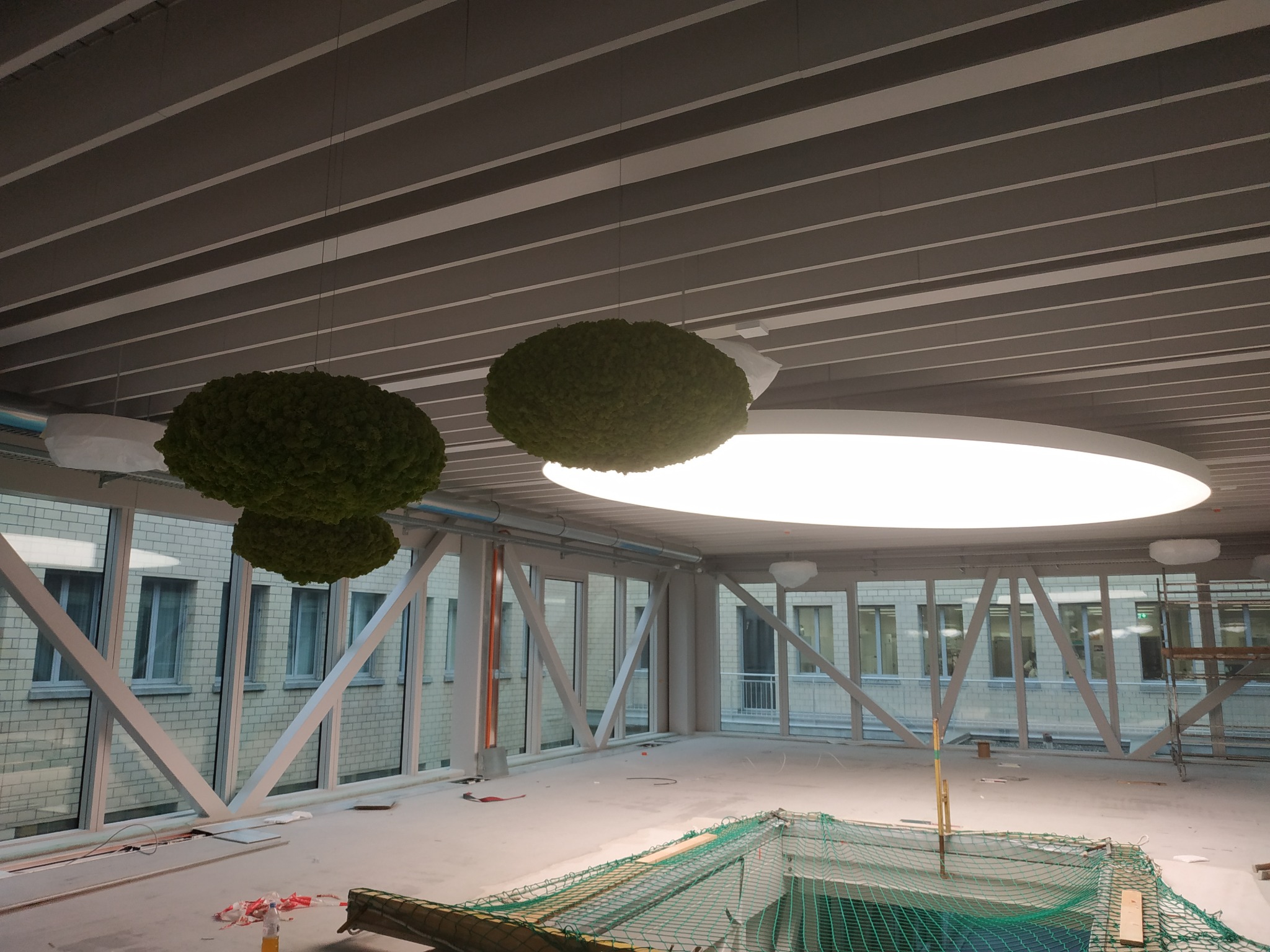 Wnętrze korytarza z przeszklonymi ścianami i podwieszonym sufitem, na którym zamontowane są okrągłe lampy i dekoracyjne kule z mchu, widoczna siatka zabezpieczająca na podłodze.