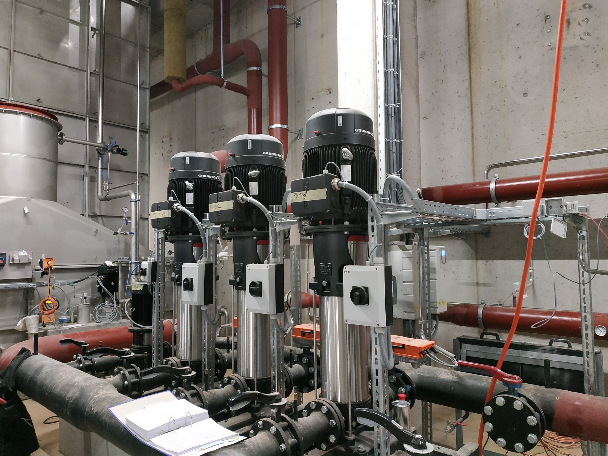 Trzy pompy Grundfos zamontowane w kotłowni, widoczne rury, zawory, przewody elektryczne i dokumentacja techniczna na podłodze