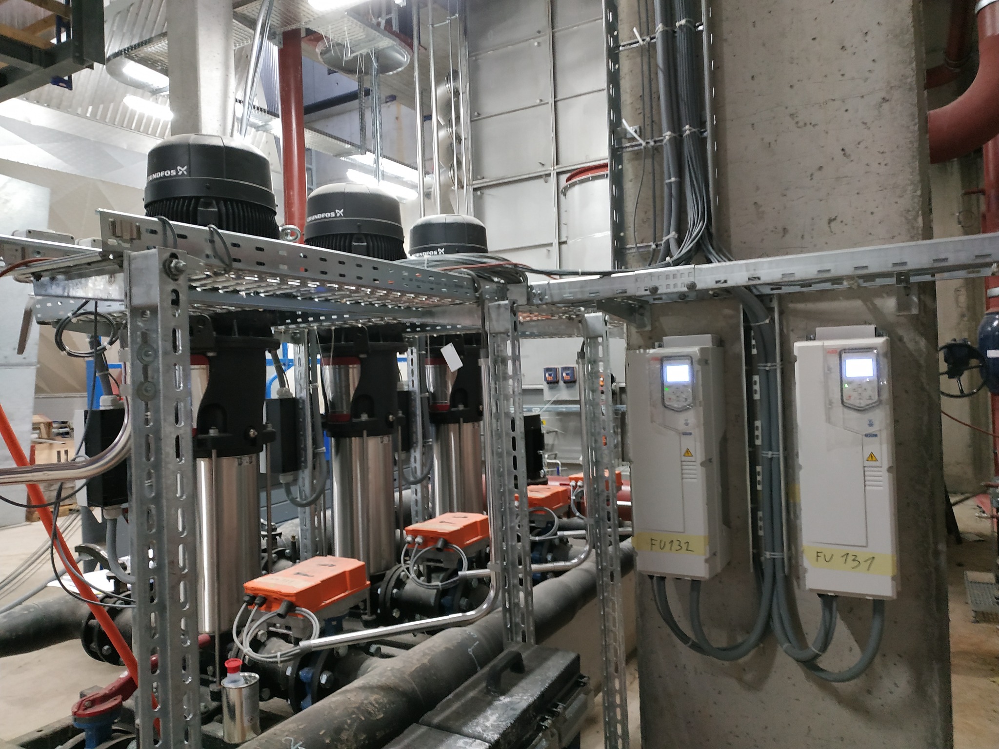 Zestaw trzech identycznych pomp Grundfos zamontowanych na metalowej konstrukcji w pomieszczeniu technicznym, z widocznymi przewodami i panelami sterowania FU132 i FU131 na betonowej ścianie.