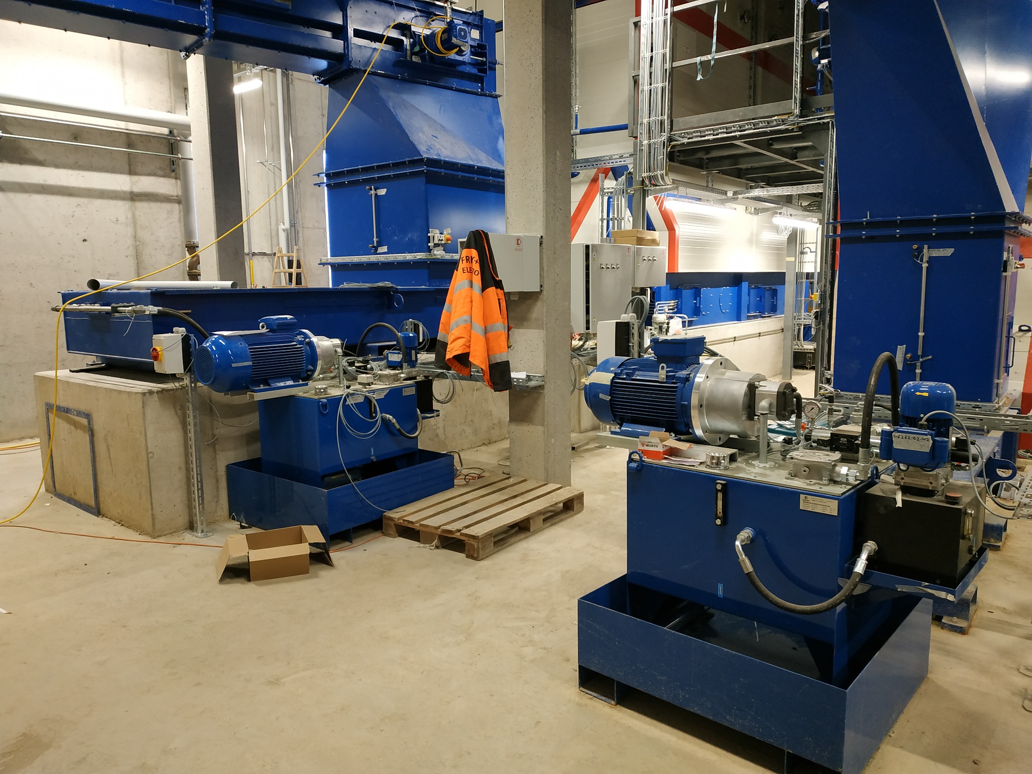 Fragment hali przemysłowej z dwoma niebieskimi agregatami hydraulicznymi, na jednym z nich wisi pomarańczowa kurtka robocza, w tle widoczne rury i instalacje pod sufitem.