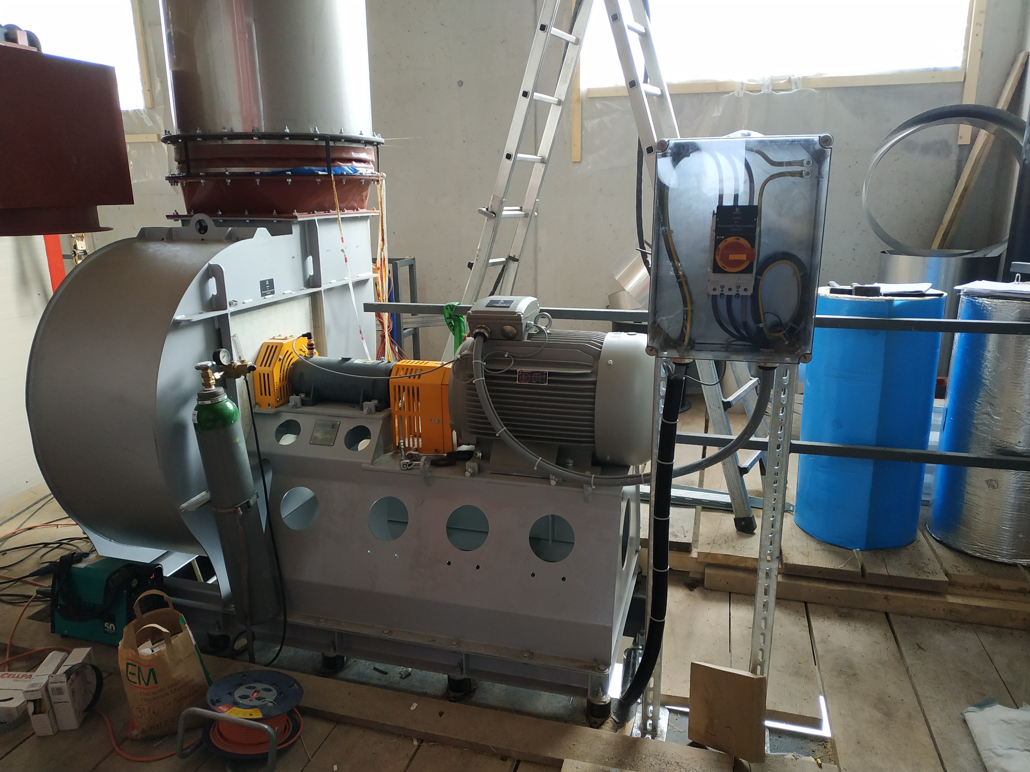 Szara przemysłowa turbina wentylacyjna z podłączonym silnikiem elektrycznym, butlą z gazem technicznym i skrzynką elektryczną w trakcie montażu, widoczna drabina i elementy konstrukcyjne budynku...