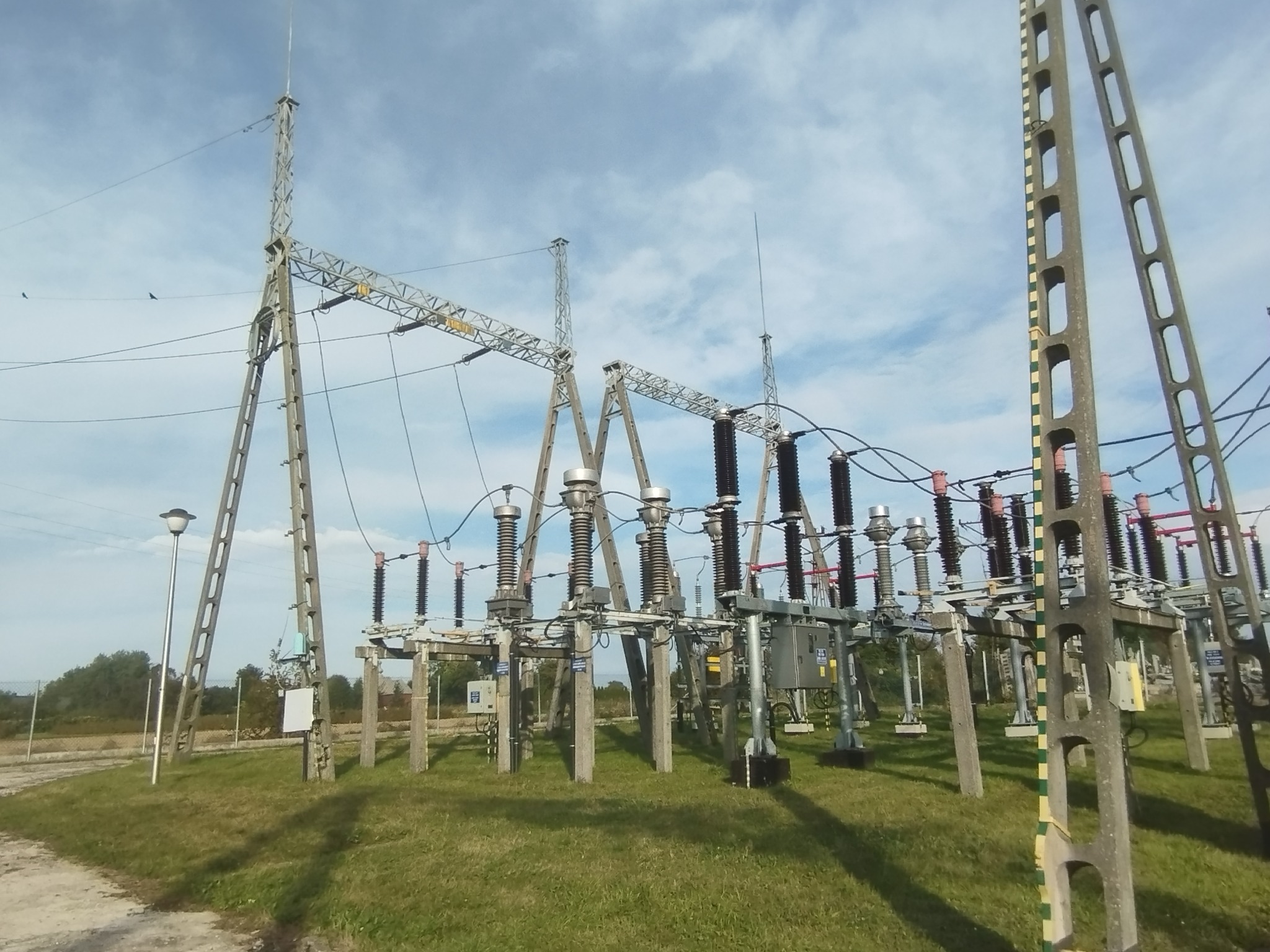 Stacja transformatorowa z betonowymi słupami kratownicowymi i izolatorami, widok z poziomu gruntu na tle błękitnego nieba z chmurami.