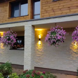 Elewacja domu jednorodzinnego z jasnego kamienia, oświetlona lampami ściennymi, udekorowana wiszącymi donicami z różowymi kwiatami.