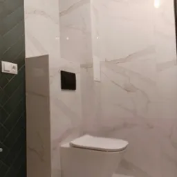 Nowoczesna łazienka z wiszącą toaletą, wykończona białymi płytkami imitującymi marmur i ciemnozielonymi płytkami ułożonymi w jodełkę, widoczny czarny przycisk spłuczki i białe gniazdko elektryczne.